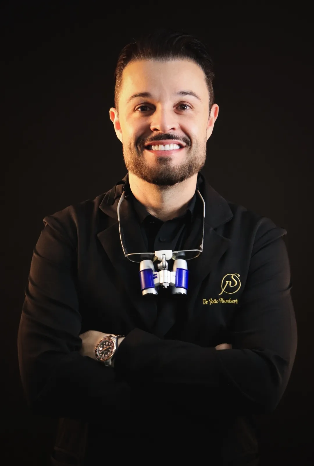 Dr. João Salgado de braços cruzados, com jaleco preto e lupa odontológica